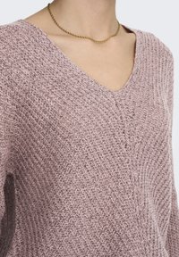 Maglione rosa a maglia con scollo a V, caratterizzato da un motivo testurizzato e una vestibilità ampia. Accompagnato da una sottile collana d'oro twistata.