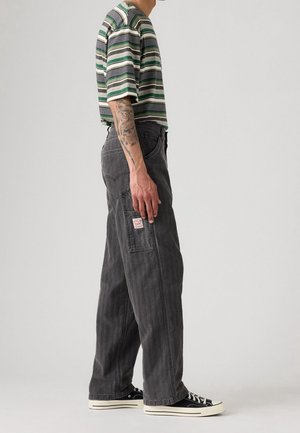 Personne portant un pantalon noir à jambes larges avec une poche plaquée, un t-shirt rayé vert et blanc, et des baskets noires, debout de profil.