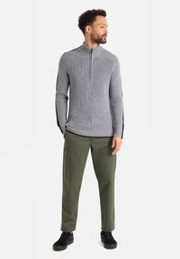 Grauer Reißverschluss-Pullover mit gerippter Textur und schwarzen Seitenelementen, kombiniert mit olivgrünen Hosen und schwarzen Sneakers, was einen kontrastierenden Look erzeugt.