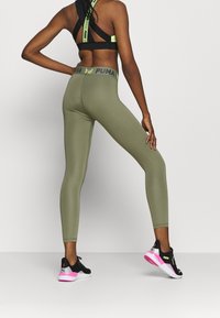 Lejki w oliwkowym kolorze o dopasowanym kroju i wysokim staniku z czarno-żółtym logo. Połączone z czarnymi sneakersami, widocznymi różowymi akcentami.