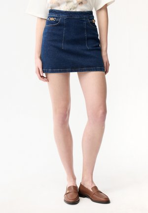 Kvinde iført en kort mørkeblå denimnederdel med dekorative gyldne detaljer og brune læderloafers, stående mod en ensfarvet baggrund.