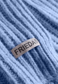 Lichtblauwe gebreide stof met diagonale ribbels; heeft een beige label met "FRIEDA" in donkere tekst gedrukt. Zachte, gestructureerde uitstraling.