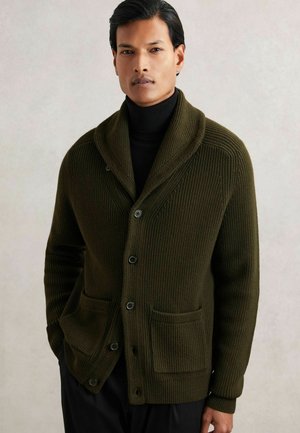 Olivgrüner gerippter Cardigan aus dickem Stoff, mit Schalkragen, vorderen Taschen und Knopfverschluss. Schmale Passform mit strukturiertem Detail.