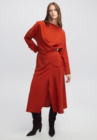 Vestido rojo de manga larga con diseño drapeado y dobladillo asimétrico, combinado con botas de gamuza negra hasta la rodilla. Tejido suave y texturizado.