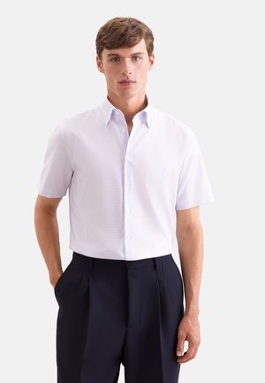 Homme portant une chemise violette claire à manches courtes avec col et un pantalon bleu marine foncé, debout avec une main dans la poche.