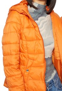 Casaco acolchoado laranja com capuz, apresentando uma textura em acolchoado e um botão de pressão prateado. Usado por cima de uma blusa cinza com padrão.