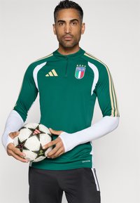 Πράσινο αθλητικό φούτερ με χρυσές λεπτομέρειες και λευκά μανίκια, που φέρει το λογότυπο "Italia" και κρατάει μια λευκή μπάλα ποδοσφαίρου με σχέδιο αστέρων.