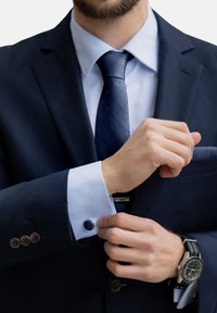 Mėlynas švarkas su šviesiai mėlynu marškiniu. Manžetėje yra apvalus mėlynas apyrankės mygtukas. Matomas laikrodis su juodu diržu.