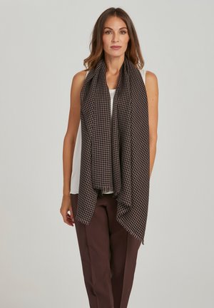 Vrouw draagt een mouwloze witte top, bruine broek en een grote zwart-bruine geruite sjaal over haar schouders, staand tegen een grijze achtergrond.