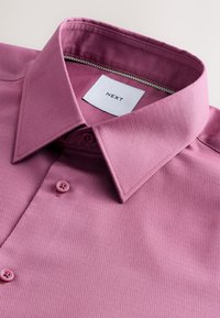 Kurzärmliges pinkfarbenes Hemd mit strukturierter Webart, ausgestattete mit einem Haifischkragen und Knopfverschluss. Etikett trägt die Aufschrift "NEXT." Die Kunststoffknöpfe sind rund.