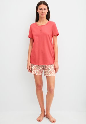 Jonge vrouw staat op blote voeten, draagt een koraalroze top met korte mouwen en korte broek met bloemenpatroon tegen een effen witte achtergrond.