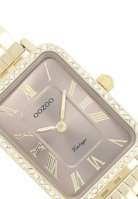 Montre rectangulaire dorée avec un cadran gris, chiffres romains, mouvement à quartz et une lunette ornée de cristaux transparents. Bracelet en métal à maillons.