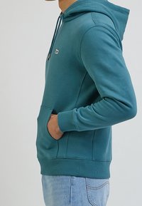 Sudadera color azul verdoso fabricada con tela suave, que incluye un bolsillo frontal tipo canguro, capucha con cordones, puños acanalados y un pequeño logo en el pecho.
