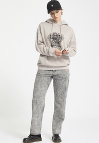 Jasno beżowa bluza z kapturem z graficznym nadrukiem przedstawiającym szkielety, w zestawieniu z szarymi dżinsami w acid wash i czarnymi butami. Czapka w kolorze czarnym dopełnia cały wygląd.