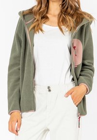 Mujer vistiendo una chaqueta de forro polar verde oliva con detalles en rosa sobre una camiseta blanca y pantalones blancos de cintura alta, con la mano derecha en el bolsillo.