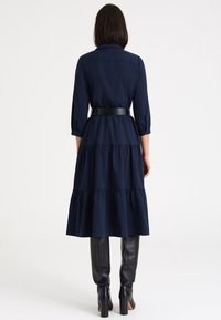Vestido camisero azul marino en capas con cintura ceñida. Presenta mangas tres cuartos y cuello con solapa. Combinado con botas negras hasta la rodilla.