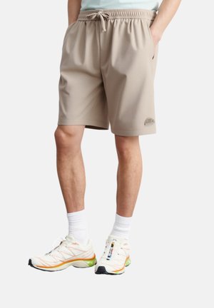 Pantaloni sportivi beige con cintura elastica, tasche laterali e un piccolo logo ricamato sull'orlo. Indossati con sneakers bianche con accenti di colore.