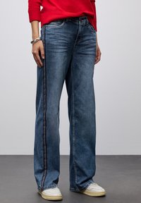 Jeans anchos y casuales de denim azul desgastado que presentan costuras laterales en contraste y un ajuste relajado, combinados con zapatillas blancas.