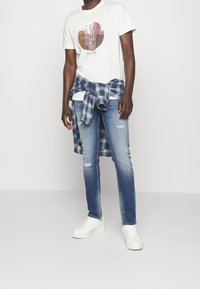 Camiseta blanca con gráfico circular, combinada con jeans desgastados de color azul claro y una camisa de cuadros azul atada en la cintura, rematada con zapatillas blancas.