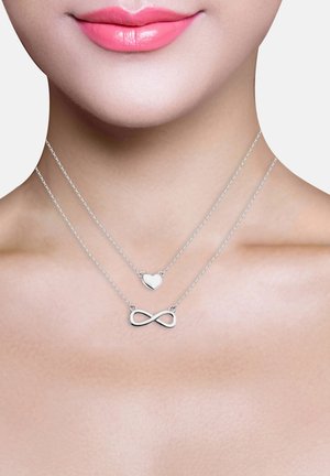 Collane a strati in argento con pendenti a forma di cuore e infinito; catene delicate; superfici lisce e lucide. Sfondo color carne.