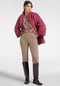 Růžový vlněný kabát s velkými knoflíky, vzorovaný hnědý svetr, kostkované béžové kalhoty a lesklé hnědé vysoké kozačky.