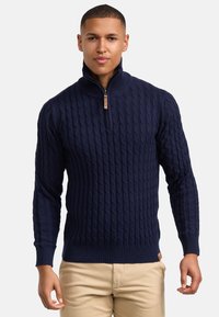 Dunkelblauer Strickpullover mit hohem Kragen, der ein strukturiertes Zopfmuster und einen Reißverschluss am Hals aufweist. Flacher Saum und gerippte Bündchen.