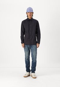 Long-sleeve black shirt with vertical stripes, light blue cap, blue jeans, and white sneakers with brown soles. Casual outfit, standing pose.