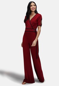 Tuta di stile uniforme rossa con scollatura a V incrociata, maniche corte, vita con cintura e rifiniture nere, caratterizzata da gambe ampie e una texture liscia.