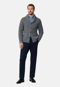 Cárdigan gris con canalé y cuello chal, cierre de botones y bolsillos delanteros, llevado sobre una camisa azul de cuello y combinado con pantalones azul marino.