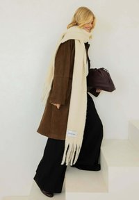 Bruine suède jas, crèmekleurige sjaal met franjes, zwarte wijde broekspijpen en een bordeauxrood tasje met trekkoord. Model die witte trappen oplopen.