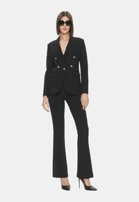Blazer nero su misura con bottoni dorati, abbinato a pantaloni attillati coordinati. La modella indossa occhiali da sole neri oversize e décolleté nere a punta.