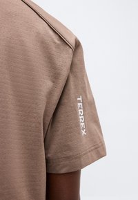 Chemise marron à manches courtes sur le bras d'une personne, présentant un texte vertical blanc "TERREX" près de l'ourlet de la manche sur un fond clair uni.