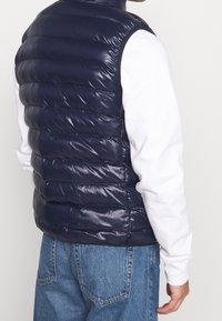 Man som bär en glänsande marinblå kviltad ärmlös väst över en vit långärmad skjorta och blå jeans, sedd bakifrån mot en ljus, enfärgad bakgrund.