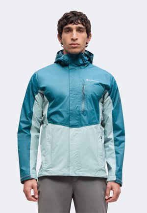 Hombre de pie con chaqueta Columbia combinada en colores azul verdoso y azul claro con capucha y bolsillos con cremallera sobre un fondo liso.