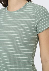 Haut rayé vert en tissu côtelé, avec des rayures verticales blanches, un col rond et de petites manches froncées.