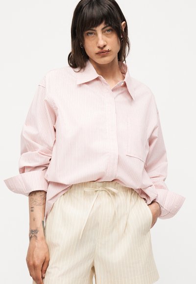 Άτομο με καστανά μαλλιά που φοράει υπερμεγέθη ριγέ ροζ πουκάμισο και μπεζ σορτς με κορδόνι, στέκεται με το ένα χέρι στην τσέπη.