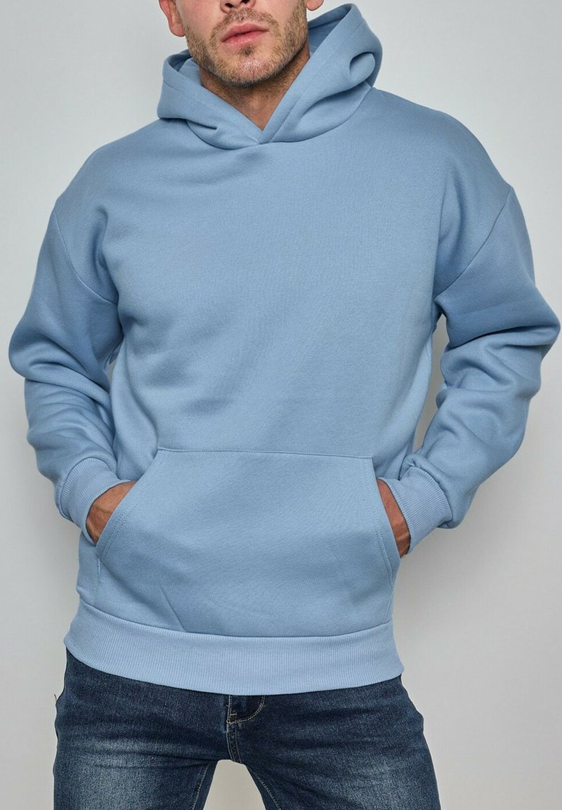 Homme portant un sweat à capuche bleu clair avec une poche frontale, les mains partiellement dans les poches, associé à un jean bleu foncé.