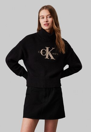 Suéter negro de punto con cuello vuelto y dobladillo acanalado, que presenta un logo beige "CK" y "Calvin Klein Jeans" en escritura por encima. Combinado con una falda de mezclilla negra.