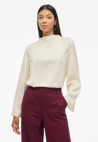 Blusa a maniche lunghe color crema con un collo alto, caratterizzata da una texture liscia e pieghe sottili, abbinata a pantaloni tailored bordeaux.