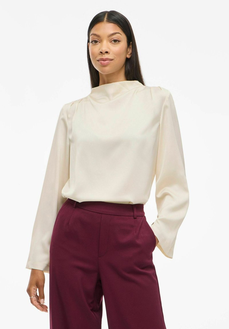 Blusa a maniche lunghe color crema con un collo alto, caratterizzata da una texture liscia e pieghe sottili, abbinata a pantaloni tailored bordeaux.