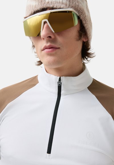 Witte zip-up jas met bruine schouderaccenten, met een hoge kraag en een subtiele logo. Grote, getinte zonnebril en een gebreide muts met strepen.