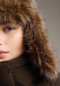 Detailní záběr osoby nosící tmavě hnědý zimní kabát s hustou, načechranou hnědou kožešinovou kapucí na jednobarevném šedém pozadí.