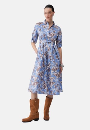 Donna che indossa un vestito midi blu a fiori con maniche 3/4 e stivali di pelle scamosciata marrone a metà polpaccio, in piedi con le mani nelle tasche su sfondo bianco.