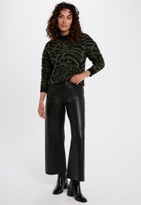 Groene trui met zwart dierenpatroon, gecombineerd met een hooggesneden, glanzend zwarte leren broek met wijde pijpen en zwarte enkelhoge laarzen.