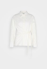 Chemise blanche à manches longues avec boutons, col et détail de lien latéral à la taille sur fond clair.