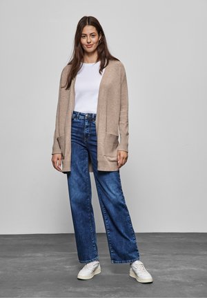 Beige gestrickte Strickjacke mit offenen Vorderteil und Taschen, kombiniert mit lockeren blauen Jeans und weißen Sneakers. Grauer Hintergrund.