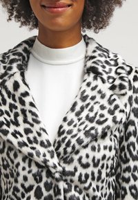 Persona vistiendo un suéter de cuello alto blanco y un abrigo de piel sintética moteado en blanco y negro con grandes solapas.