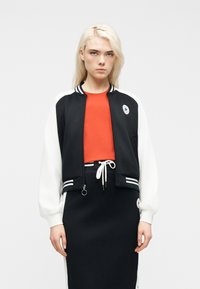 Νεαρή γυναίκα με μακριά ξανθά μαλλιά, φορώντας μαύρο και άσπρο varsity μπουφάν, κόκκινο πουκάμισο και μαύρη φούστα με κορδόνι, σε λευκό φόντο.