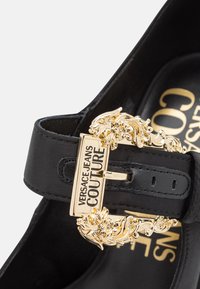 Czarne skórzane buty z dekoracyjnym złotym zapięciem z kwiatowym wzorem i tłoczonym logo. Gładka tekstura z eleganckim wykończeniem.
