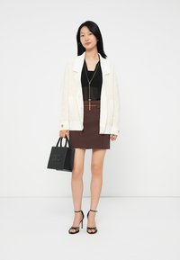 Woman wearing white textured cardigan, black sheer top, brown skirt, black strappy heels, holding black handbag with gold chain necklace.